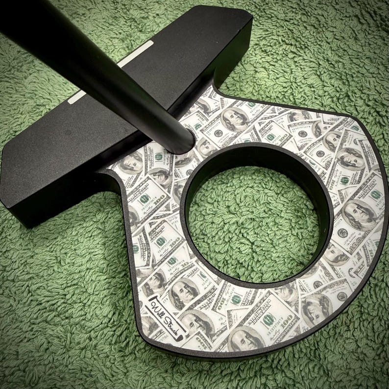 Puede incluir: Putter de golf negro con un dise&ntilde;o tem&aacute;tico de dinero. La cabeza del putter presenta un patr&oacute;n de billetes de cien d&oacute;lares. El putter tiene un eje negro y una abertura circular en la cabeza. Las palabras "Will Struck" est&aacute;n impresas en el putter.