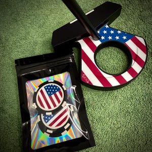 May include: A black golf putter with a patriotic American flag design on the club head. The putter features red and white stripes and a blue field of stars. Two golf ball markers with the same design are in a black bag.