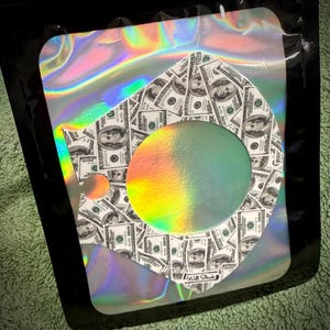 Puede incluir: Una bolsa hologr&aacute;fica negra con un dise&ntilde;o tem&aacute;tico de dinero. El dise&ntilde;o presenta una forma de diamante con un c&iacute;rculo recortado, lleno de im&aacute;genes de billetes de 100 $. El c&iacute;rculo y los bordes de la bolsa tienen un brillo de arco&iacute;ris. El texto "Will Stick" es visible.