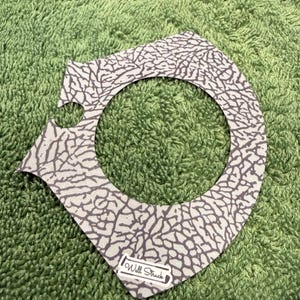 Pu&ograve; includere: Un accessorio grigio e bianco a fantasia con un ritaglio circolare. Il motivo ricorda la pelle di elefante. Il testo "Will Struck" &egrave; stampato su una piccola etichetta bianca nella parte inferiore dell'accessorio.