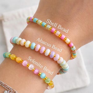 Puede incluir: Tres pulseras de cuentas en colores pastel se muestran en una muñeca. Las pulseras están etiquetadas como "Shell Bead", "Abacus Bead" y "Selenite Bead". Cada pulsera presenta un estilo de cuentas y una combinación de colores diferente, con espaciadores dorados.