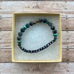 May include: A beaded bracelet with the words "BE TRANSFORMED" spelled out in black and white letter beads. The bracelet features gray and turquoise beads, along with gold-colored metal accents. The bracelet is presented in a small, light-yellow box.