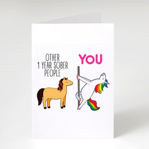 Pu&ograve; includere: Un biglietto d'auguri bianco con un cavallo marrone chiaro disegnato con il testo OTHER 1 YEAR SOBER PEOPLE e un unicorno bianco con criniera arcobaleno che danza su un palo con il testo rosa YOU, per celebrare un anno di sobriet&agrave;.