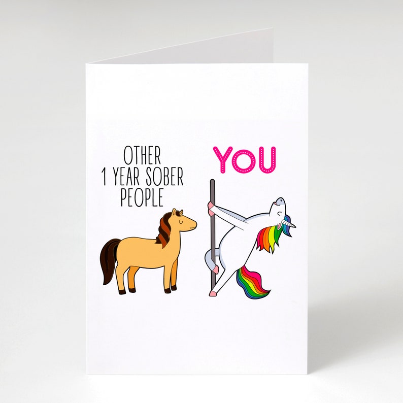 Pu&ograve; includere: Un biglietto d'auguri bianco con un cavallo marrone chiaro disegnato con il testo OTHER 1 YEAR SOBER PEOPLE e un unicorno bianco con criniera arcobaleno che danza su un palo con il testo rosa YOU, per celebrare un anno di sobriet&agrave;.