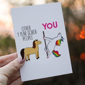 Pu&ograve; includere: Un biglietto d'auguri bianco con un'illustrazione a cartoni animati di un cavallo marrone etichettato OTHER 1 YEAR SOBER PEOPLE e un unicorno bianco con criniera e coda arcobaleno che balla su un palo etichettato YOU in testo rosa.