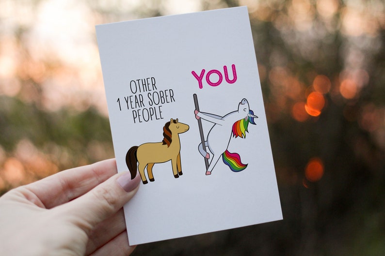 Pu&ograve; includere: Un biglietto d'auguri bianco con un'illustrazione a cartoni animati di un cavallo marrone etichettato OTHER 1 YEAR SOBER PEOPLE e un unicorno bianco con criniera e coda arcobaleno che balla su un palo etichettato YOU in testo rosa.