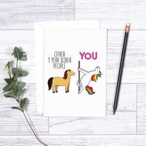 Pu&ograve; includere: Un biglietto d'auguri bianco con un cavallo color marrone chiaro in stile cartoon accanto al testo OTHER 1 YEAR SOBER PEOPLE e un unicorno con criniera arcobaleno che danza su un palo accanto al testo rosa YOU, con busta bianca.