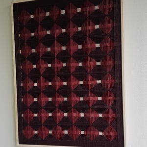 May include: Framed wall art with a geometric design. The pattern features dark burgundy and red diamond shapes, with small white squares at the intersections. The artwork is framed in light-coloured wood.