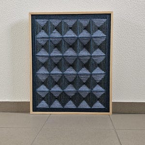 May include: A rectangular wall art piece featuring a grid of dark blue and black yarn wrapped into three-dimensional pyramid shapes. The artwork is encased in a light-colored wooden frame, creating a textured geometric pattern across the surface.