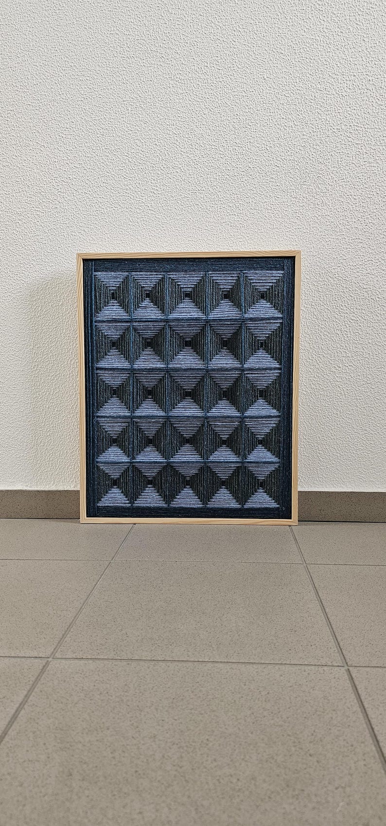 May include: A rectangular wall art piece featuring a grid of dark blue and black yarn wrapped into three-dimensional pyramid shapes. The artwork is encased in a light-colored wooden frame, creating a textured geometric pattern across the surface.