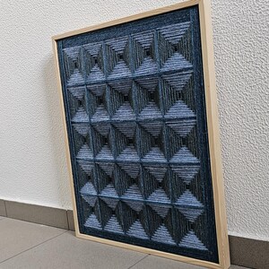 May include: A rectangular wall art piece featuring a grid of dark blue and black yarn wrapped around pins to create a three-dimensional geometric pyramid pattern, all encased within a light-colored wooden frame with a visible grain texture.