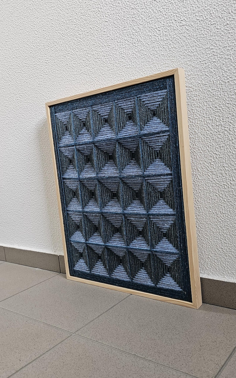 May include: A rectangular wall art piece featuring a grid of dark blue and black yarn wrapped around pins to create a three-dimensional geometric pyramid pattern, all encased within a light-colored wooden frame with a visible grain texture.