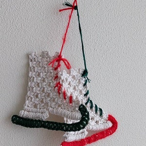 May include: White crocheted ice skate ornament with green and red string accents. The ornament is hanging from a white wall.