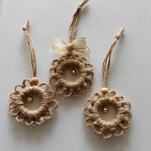 May include: Three handcrafted jute ornaments with a floral design. Each ornament features a small gold bell in the centre and a wooden bead at the top. One ornament has a gold bow. The ornaments are suspended by jute twine.