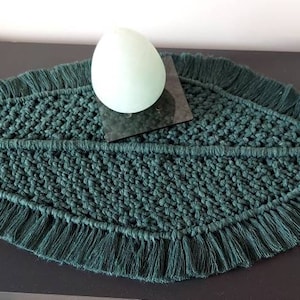 May include: A dark green leaf-shaped macrame table runner with fringe around the edges. A white egg-shaped object is sitting on top of the runner.