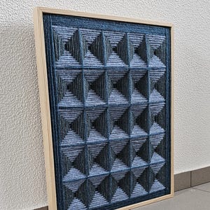 May include: A rectangular wall art piece featuring a grid of raised, pyramid-shaped structures created from dark blue and light blue yarn. The geometric design is encased in a light-colored wooden frame, showcasing a textured, layered appearance.