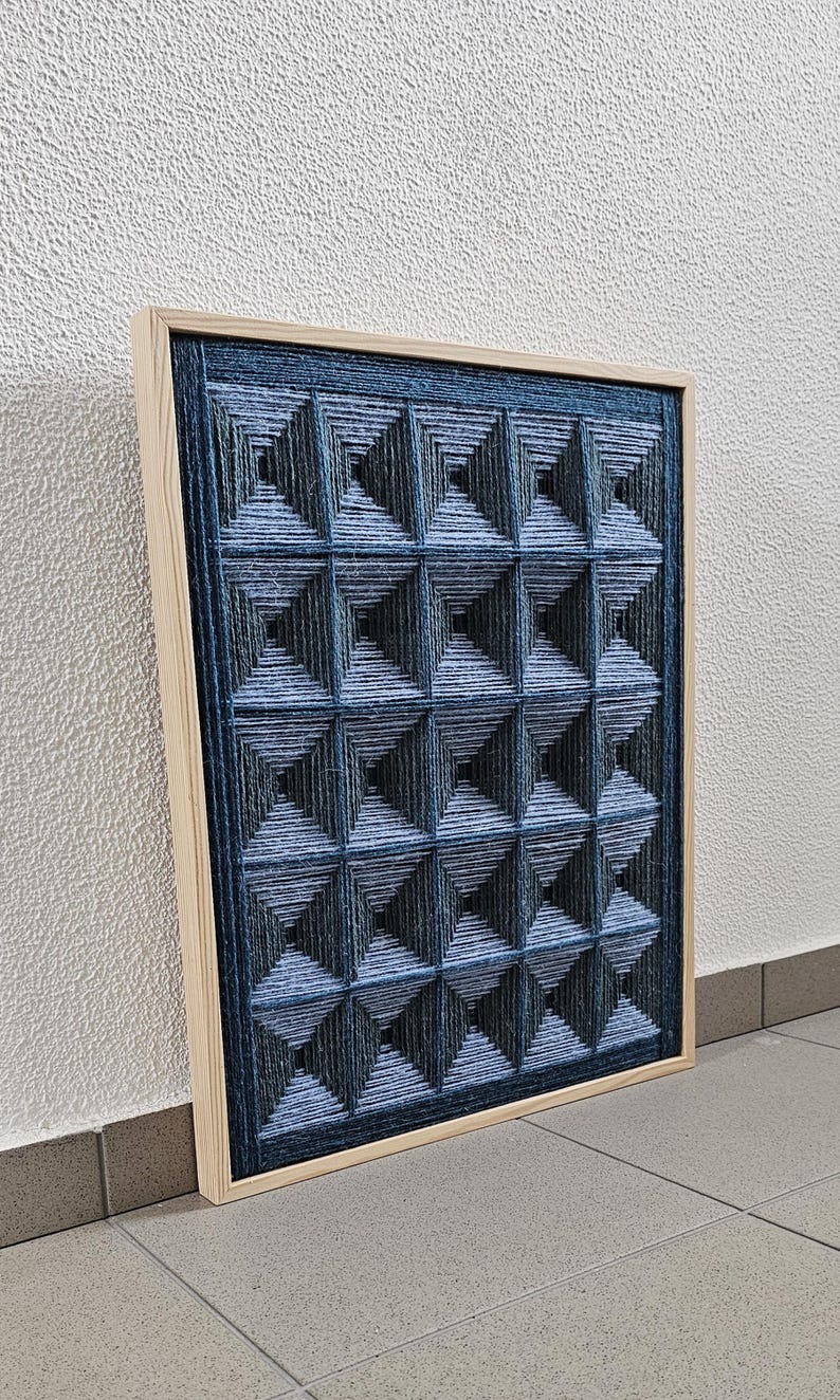 May include: A rectangular wall art piece featuring a grid of raised, pyramid-shaped structures created from dark blue and light blue yarn. The geometric design is encased in a light-colored wooden frame, showcasing a textured, layered appearance.