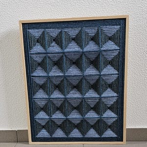 May include: A square wall art piece featuring a grid of sixteen pyramid shapes created with dark blue and black yarn wrapped around nails. The geometric design is encased in a light-colored wooden frame, showcasing a textured, three-dimensional pattern.