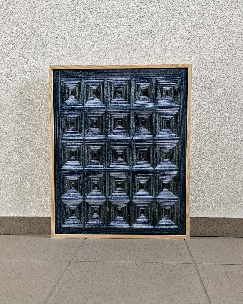 May include: A rectangular wall art piece featuring a grid of sixteen pyramid-shaped structures created with dark blue and black yarn. The yarn is wrapped tightly to form geometric patterns within a light-colored wooden frame, creating a textured effect.