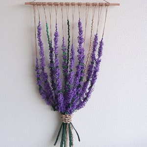 May include: A wall hanging made of yarn in the shape of lavender flowers. The flowers are purple and green and are hanging from a wooden dowel. The dowel is attached to a string that is hanging from the ceiling.