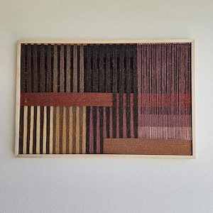 May include: Abstract fibre art in a wooden frame. The artwork features vertical and horizontal lines in shades of brown, red, yellow, and black. The woven design creates a textured, geometric pattern, suitable for home decor.