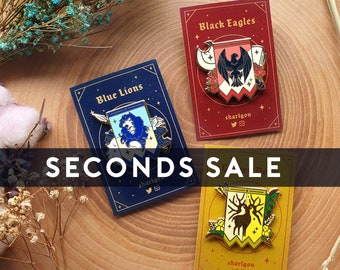 May include: Three enamel pins featuring crests from the Fire Emblem: Three Houses video game. The pins are on a wooden background with flowers and wheat. The crests are for the Blue Lions, Black Eagles, and Golden Deer houses. The pins are for sale and are marked "Seconds Sale".