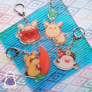 Puede incluir: Cuatro llaveros de personajes coloridos con anillos de llaves plateados. Los llaveros presentan un conejo blanco con detalles rojos, un conejo blanco con un trébol, un mapache amarillo con un chile rojo y un fantasma blanco con un sombrero rojo y flores.