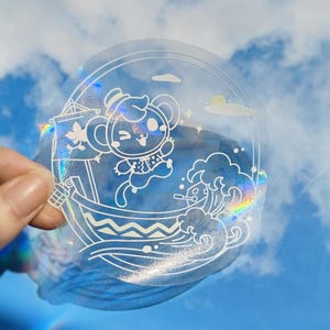 Pode incluir: Um adesivo transparente iridescente com um contorno branco de um animal de desenho animado usando um chapéu e navegando em um barco com ondas. O adesivo tem um acabamento holográfico.
