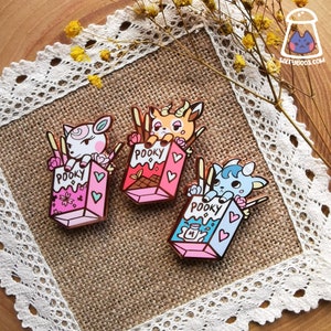 May include: Three enamel pins featuring cartoon animal characters sitting inside a box of Pocky snacks. The pins are gold-toned and have a glossy finish. The characters are a white sheep, a brown deer, and a blue cat.