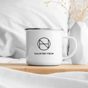 May include: White enamel mug with a silver rim, featuring a black geometric logo and the text "CALIETNE ITECH". The mug is placed on a wooden surface, with a white vase and dried flowers in the background. A simple, modern design.