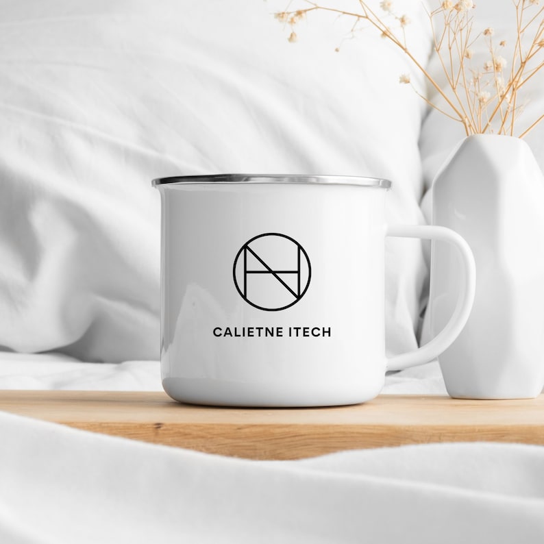 May include: White enamel mug with a silver rim, featuring a black geometric logo and the text "CALIETNE ITECH". The mug is placed on a wooden surface, with a white vase and dried flowers in the background. A simple, modern design.