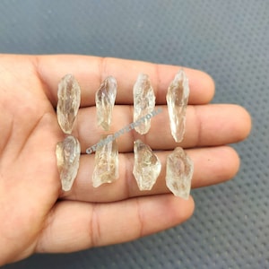 May include: Eight clear quartz crystal points, also known as rock crystals, are displayed on a hand. The crystals are translucent and have a slight yellow hue. The crystals are all different sizes and shapes.