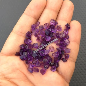 May include: A close-up image of a handful of small, purple amethyst crystals. The crystals are rough and have a natural, unpolished look. The crystals are in various shapes and sizes.