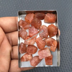K&ouml;nnte beinhalten: Ein kleiner Metallbeh&auml;lter gef&uuml;llt mit rohen, orange-roten Sunstone-Kristallen. Die Kristalle sind in verschiedenen Gr&ouml;&szlig;en und Formen.