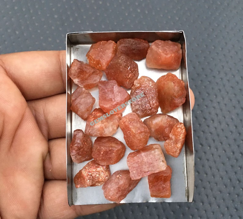 K&ouml;nnte beinhalten: Ein kleiner Metallbeh&auml;lter gef&uuml;llt mit rohen, orange-roten Sunstone-Kristallen. Die Kristalle sind in verschiedenen Gr&ouml;&szlig;en und Formen.