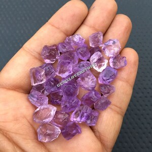 May include: A collection of small, rough-cut amethyst crystals in various shades of purple. The crystals are loose and scattered in a hand.