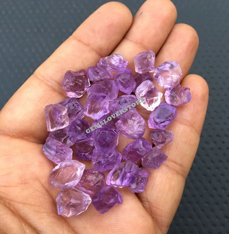 May include: A collection of small, rough-cut amethyst crystals in various shades of purple. The crystals are loose and scattered in a hand.