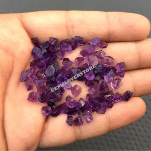 May include: A collection of small, rough amethyst crystals in various shades of purple. The crystals are scattered on a hand, showcasing their natural beauty and unique shapes.