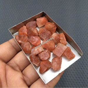 K&ouml;nnte beinhalten: Nahaufnahme eines kleinen Metalltabletts gef&uuml;llt mit rohen, orange-roten Sunstone-Kristallen. Die Kristalle sind in verschiedenen Gr&ouml;&szlig;en und Formen, einige haben einen leicht metallischen Glanz.