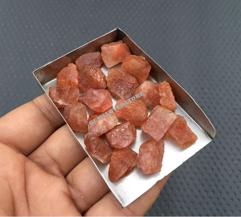 K&ouml;nnte beinhalten: Nahaufnahme eines kleinen Metalltabletts gef&uuml;llt mit rohen, orange-roten Sunstone-Kristallen. Die Kristalle sind in verschiedenen Gr&ouml;&szlig;en und Formen, einige haben einen leicht metallischen Glanz.