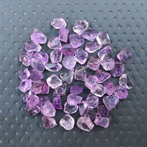 May include: A collection of approximately 50 small, rough-cut amethyst crystals. The crystals are a light purple color and have a variety of shapes and sizes.