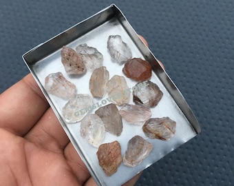 May include: A collection of raw, clear quartz crystals with red and brown inclusions. The crystals are in a small metal tray.