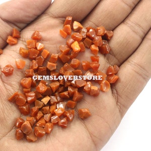 May include: A close-up of a hand holding a pile of small, irregularly shaped, orange-red gemstones. The gemstones are all about the same size and have a shiny, polished finish.