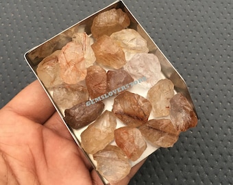May include: A collection of raw, translucent, red and brown rutilated quartz crystals in a metal tray. The crystals are rough and have a natural, unpolished appearance.