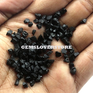 May include: A hand holding a pile of black gemstone chips. The chips are small and have a rough, irregular shape. The text "GEMSLOVERSTORE" is visible in the image.