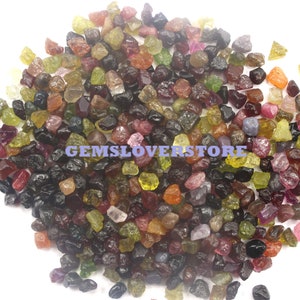 Puede incluir: Un montón grande de piedras preciosas en bruto de colores, incluyendo verde, rojo, rosa, azul y negro. Las piedras preciosas son pequeñas e irregulares en forma, y están esparcidas sobre una superficie blanca. GEMSLOVERSTORE está impreso en la imagen.