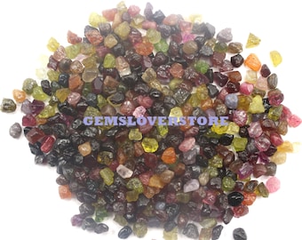 Könnte beinhalten: Ein großer Haufen bunter, roher Edelsteine, darunter grüne, rote, rosa, blaue und schwarze. Die Edelsteine sind klein und unregelmäßig geformt und auf einer weißen Oberfläche verstreut. GEMSLOVERSTORE ist auf dem Bild gedruckt.