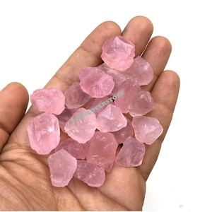 Può includere: Una collezione di cristalli di quarzo rosa, grezzi e levigati, in una mano. I cristalli sono di diverse dimensioni e forme.