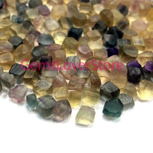 May include: A collection of small, faceted, multi-colored gemstones. The gemstones are mostly shades of green, brown, and yellow, with some purple accents. The gemstones are irregularly shaped and have a rough, natural finish.