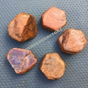 Może przedstawiać: Pięć surowych, nieoszlifowanych czerwonych kamieni szlachetnych. Kamienie szlachetne są w różnych odcieniach czerwieni i brązu, niektóre mają fioletowy odcień. Kamienie szlachetne są wszystkie nieregularne i mają naturalne, nieoszlifowane wykończenie.